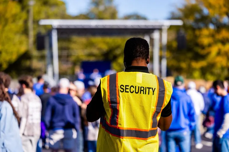 Security steward seen from behind in a yellow hi-vis vest reading ‘Security’, standing among the crowd at a daytime outdoor event, in front of an empty stage structure