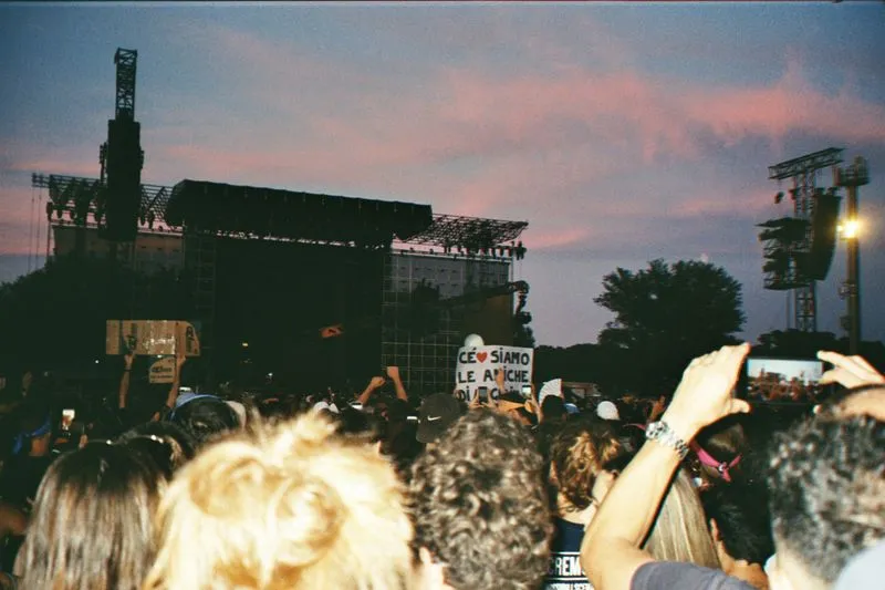 Crowd at an open-air festival at sunset, a lit stage in the distance and signs held up in the crowd
