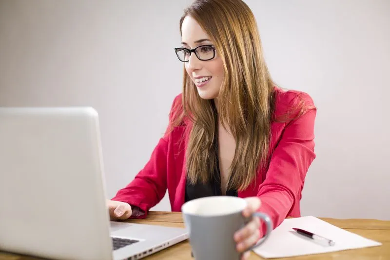 Young smiling woman sitting at a laptop, a coffee mug in hand next to the keyboard