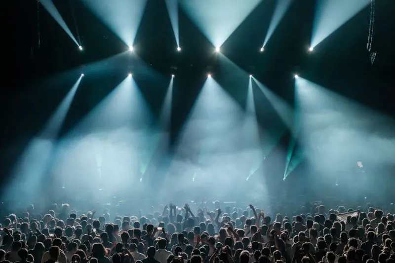 Packed crowd at a gig, arms raised towards a stage crossed by bluish light beams and haze