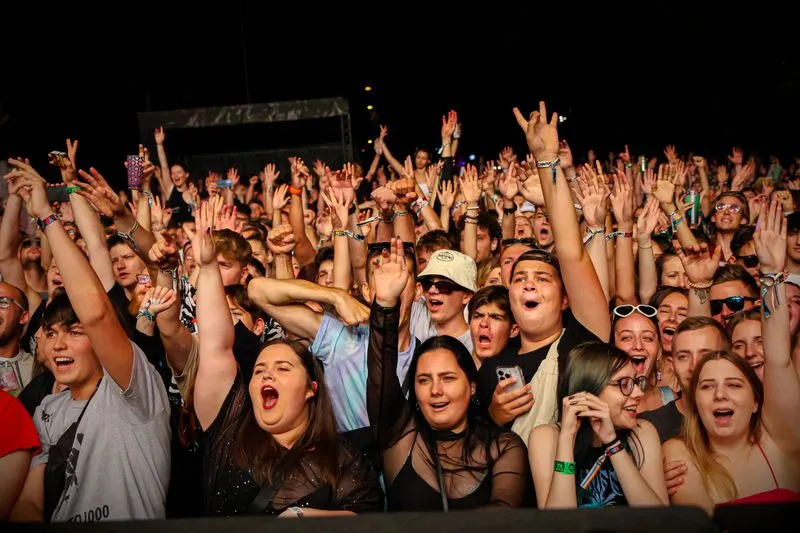 Dense crowd at a night-time gig, seen from the stage, arms raised and faces turned towards the band, a few phones held up