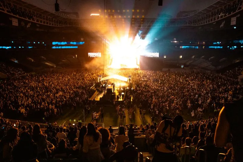 Stadium packed with fans at night, spotlights sweeping above a large stage and giant screens lit up