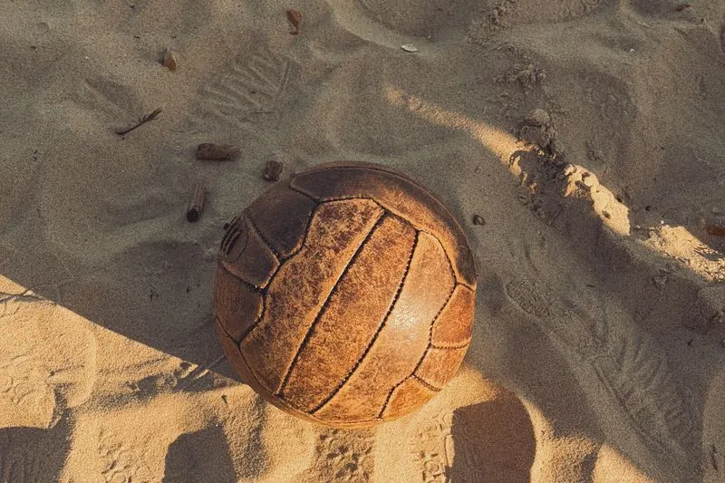 Worn brown leather football with long stitched panels, resting in the sand