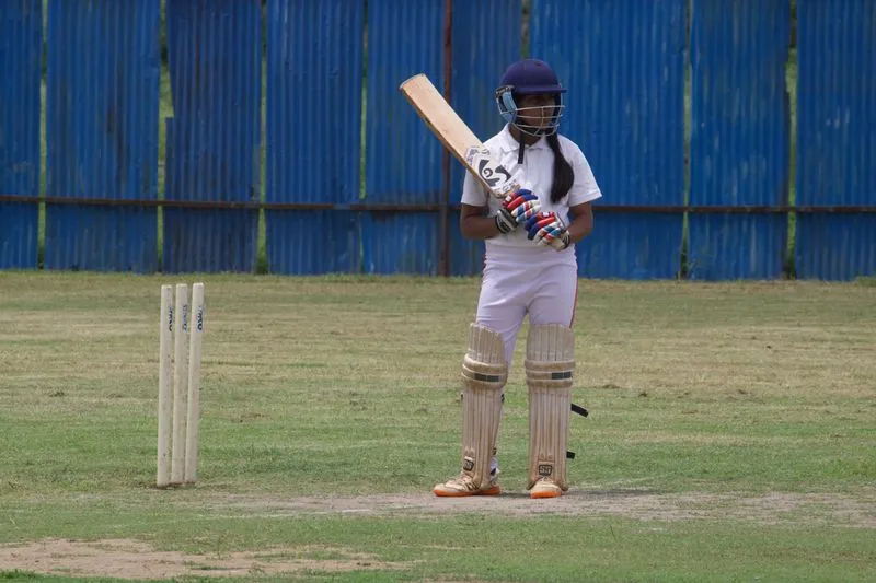 Young female cricketer in whites, helmet and pads, bat raised on her shoulder beside the stumps on a grass ground