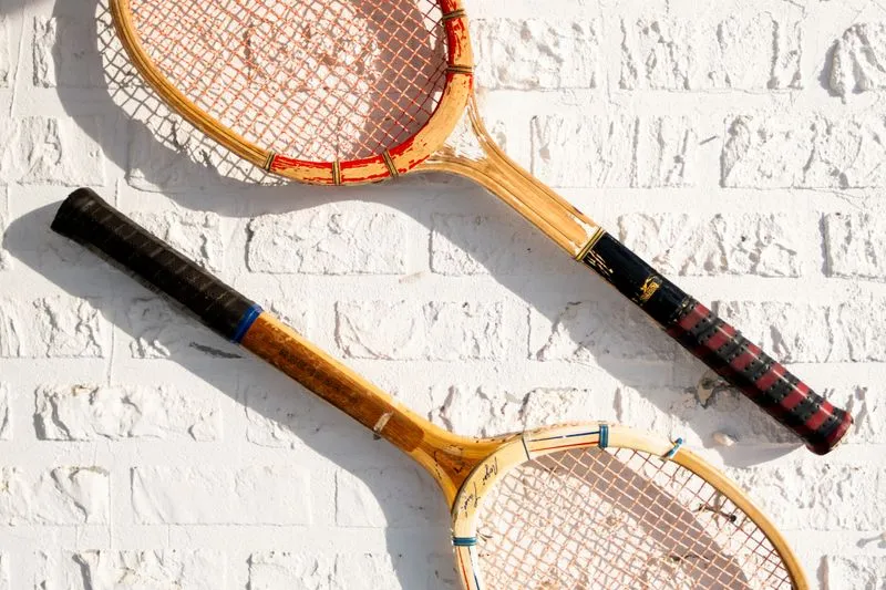 Two old wooden tennis rackets with wrapped grips, hanging in the sun on a white-painted brick wall