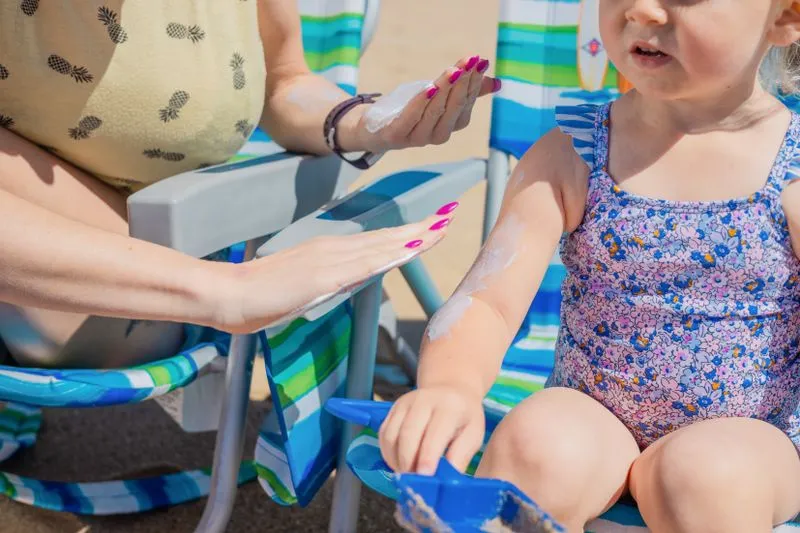 An adult spreading a thick white sunscreen on a child's arm at the beach