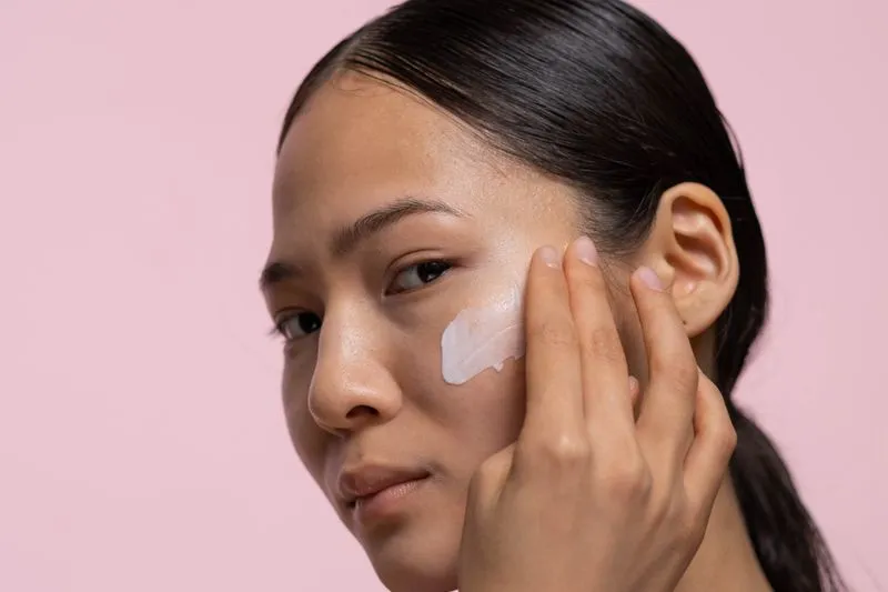 Young woman dabbing a stroke of white cream onto her cheekbone with her fingertips, against a pink background