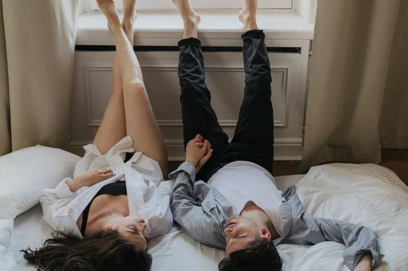 A couple lying on their backs on a bed, legs raised against a window ledge, holding hands