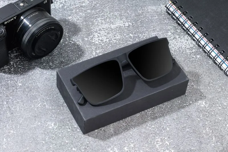 Black sunglasses resting on their open case, next to a camera, on a stone surface