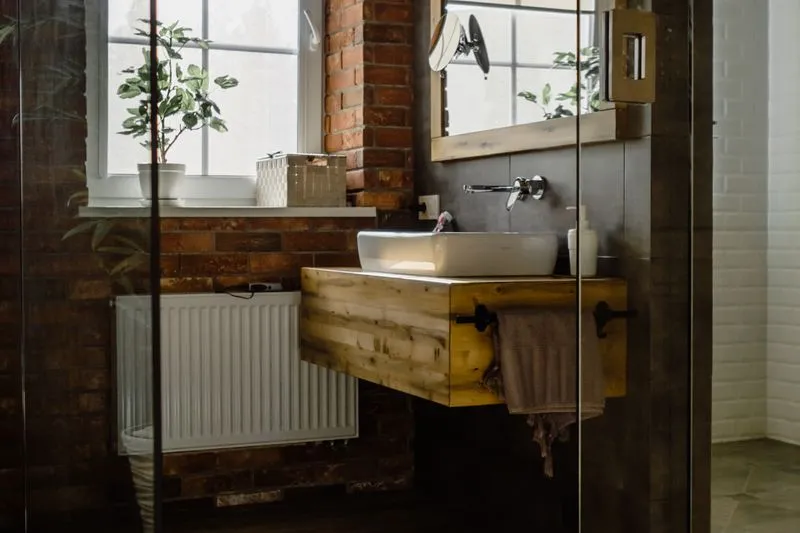 Green potted plant on a bathroom windowsill, beside a basin on a wooden vanity and a brick wall