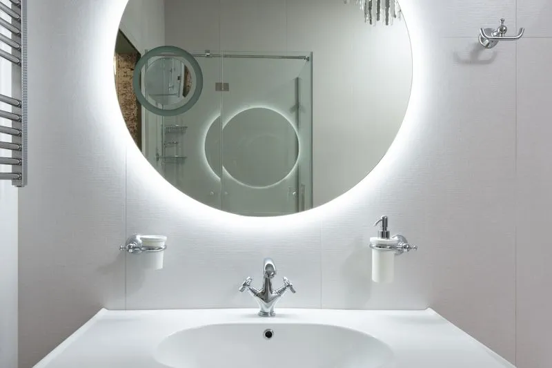 White basin under a large round backlit mirror, with a glass shower reflected in it
