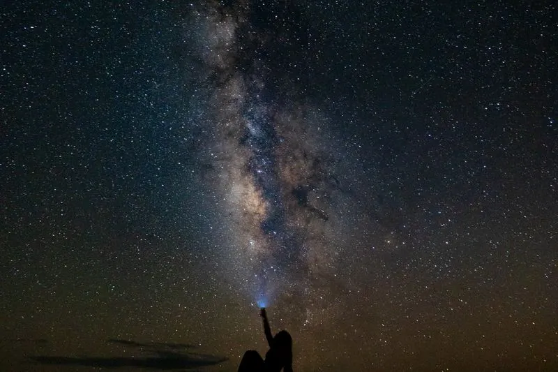 Silhouette of a person sitting outside at night, arm raised towards a starry sky crossed by the Milky Way