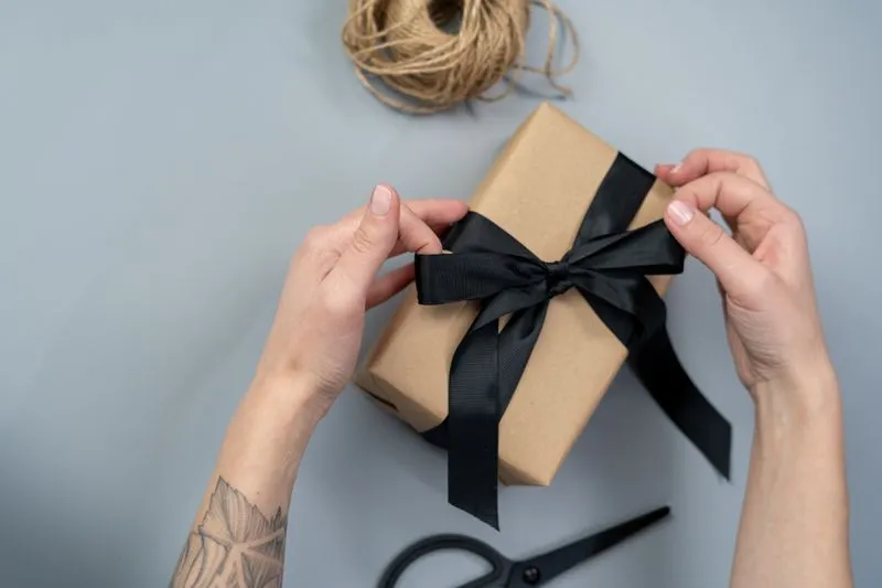 Hands tying a black ribbon around a small gift wrapped in kraft paper, a spool of twine beside it