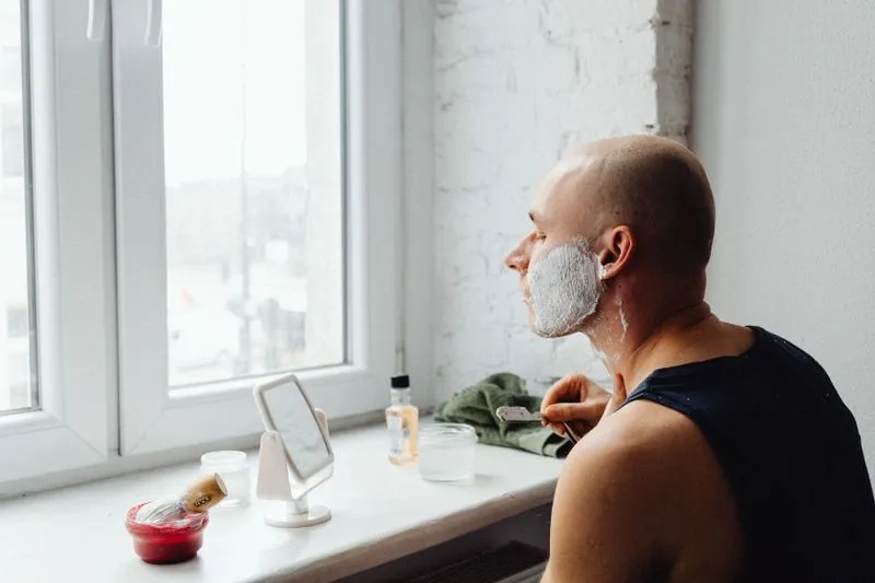 Man in a vest shaving by a window, shaving foam on his face, using a small mirror propped on the sill