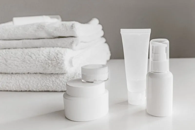 Stacked cream jars, a tube and a pump bottle, all white, on a bathroom counter with folded towels behind