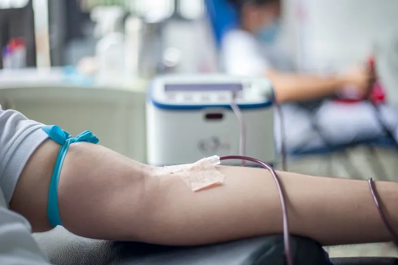 An arm connected by a tube to a collection machine, with another donor visible in the background