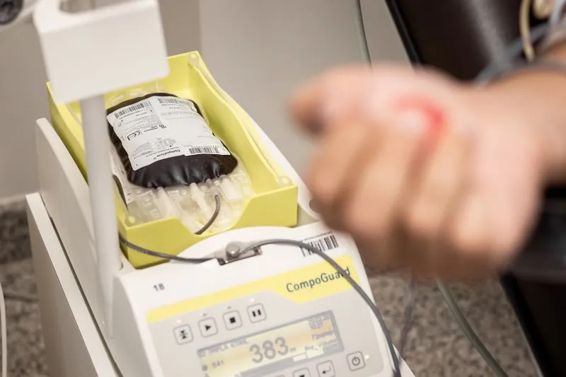 A blood bag inside a weighing and mixing device, with a hand bearing a fresh puncture mark blurred in the foreground
