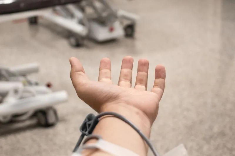 An open hand and forearm with a grey tube looped around the wrist, medical equipment on wheels in the background