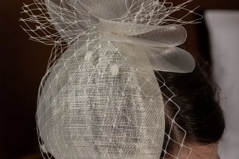 White sinamay headpiece with a net veil, worn to one side on dark hair