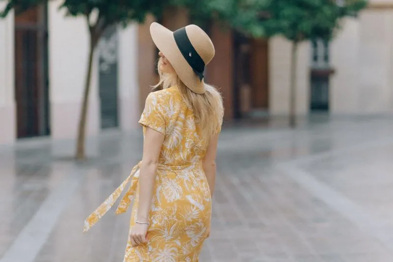 Woman seen from behind in a yellow printed dress and a straw hat with a black ribbon, on a cobbled square