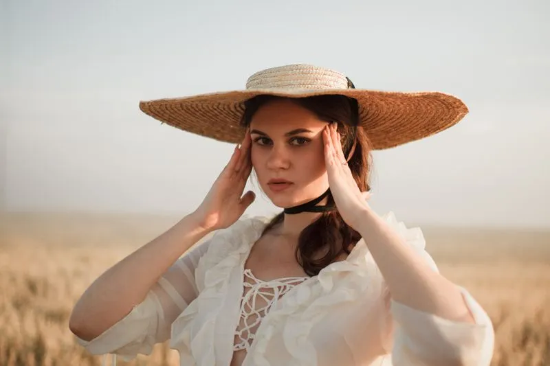 Woman in a white ruffled dress and wide-brimmed straw hat, hands framing her face, standing in a wheat field
