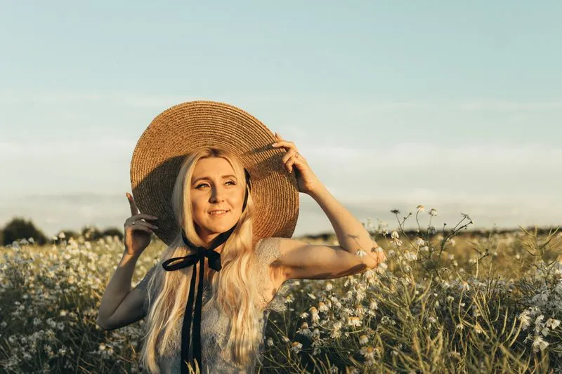 Smiling woman in a wide-brimmed straw hat with a black ribbon, holding its brim, in a wildflower meadow
