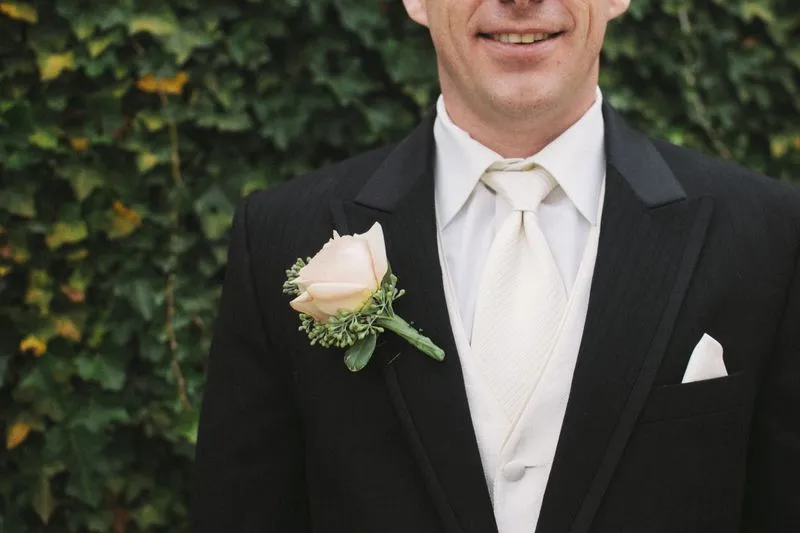 Close-up of a black suit with a cream waistcoat and tie and a pale pink rose buttonhole, against an ivy wall