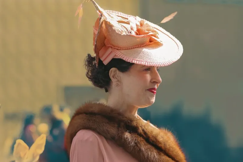 Woman in profile in a pink outfit, a large pink straw hat trimmed with feathers and a fur stole