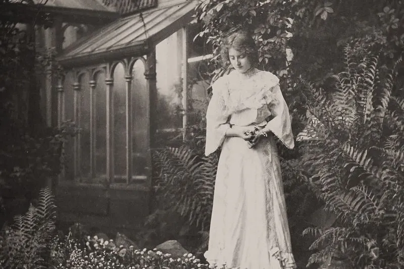 Vintage sepia photograph of a woman in a long white ruffled dress with flared sleeves, standing among ferns in front of a glasshouse
