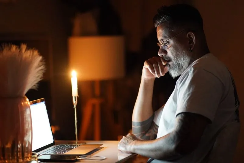 Man alone at a laptop with a candle beside him, late in the evening in a dark room