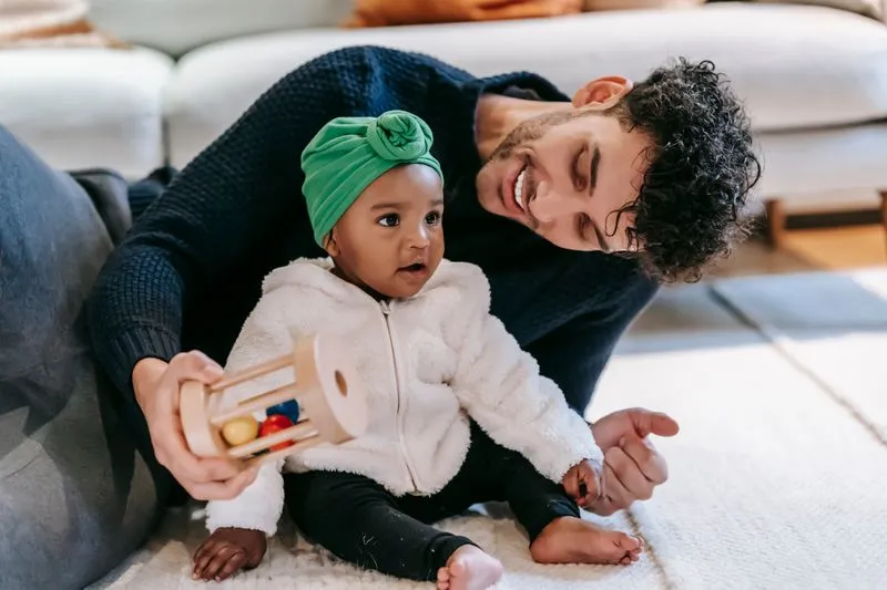 Father lying on the floor, holding a toy, smiling at his baby sitting on a rug