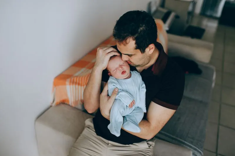 Father sitting and cradling his swaddled newborn, kissing his forehead