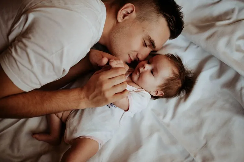 Young father lying on a bed, eyes closed, his face pressed close to his baby's