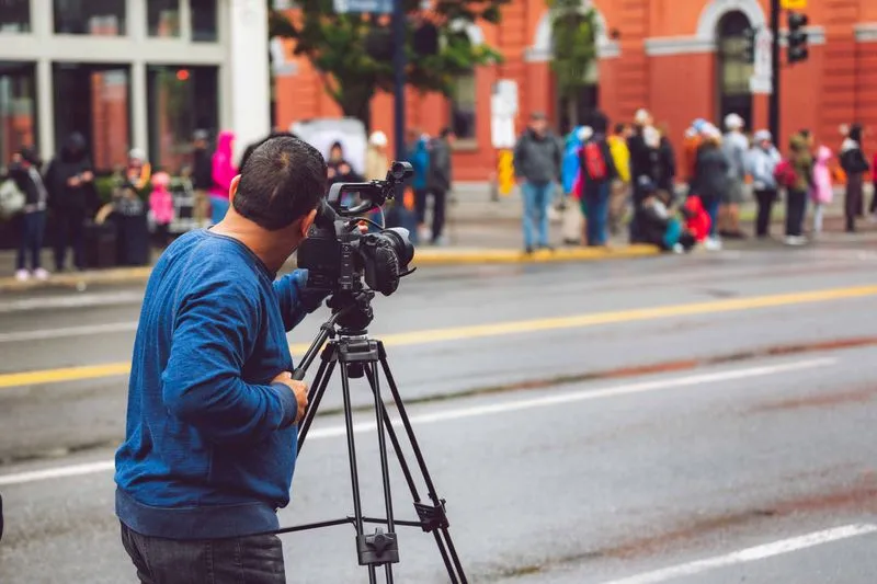 A camera operator filming a street from a tripod, with people in raincoats gathered on the pavement opposite
