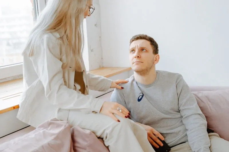 A woman standing by a sofa rests her hand on her partner's shoulder as he looks up at her, worried