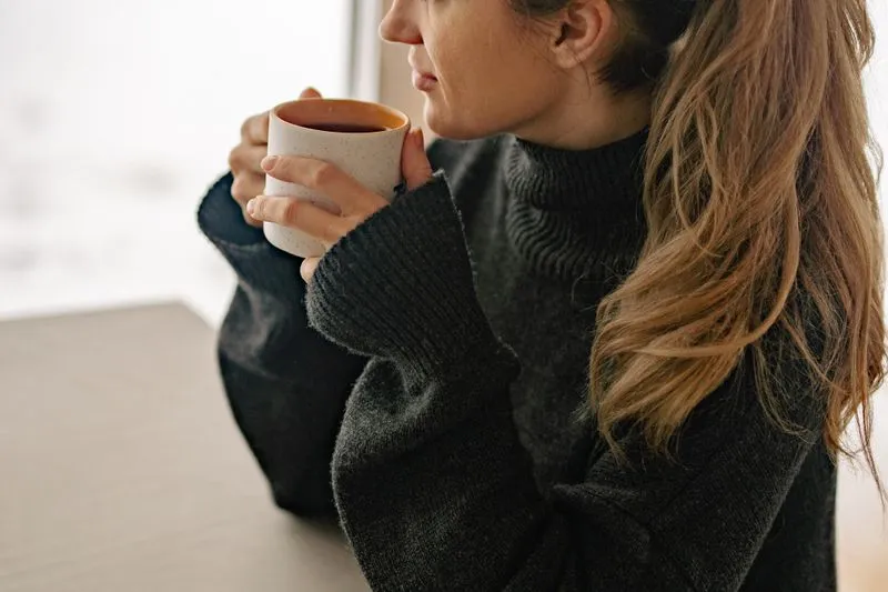 A woman in a dark roll-neck jumper, holding a mug and turned towards a window