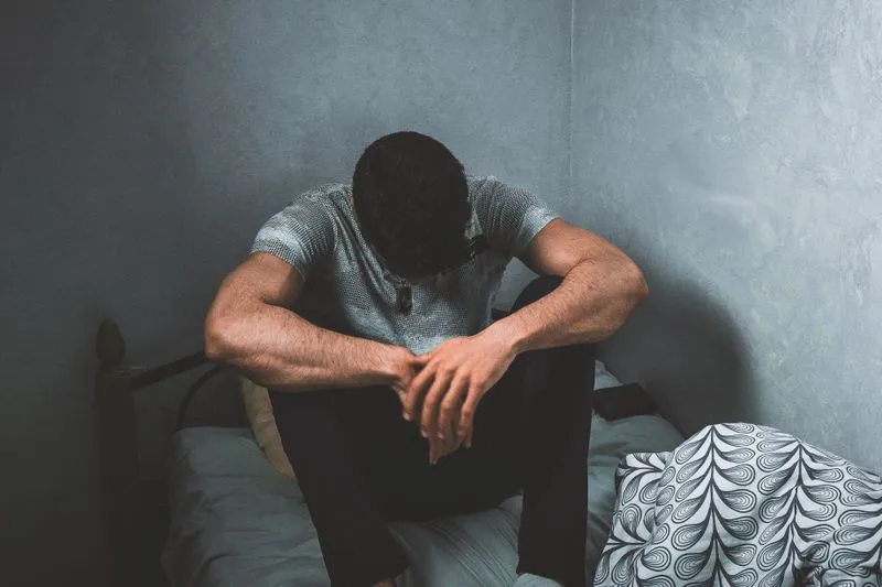 A man sitting on the edge of a bed, elbows on his knees, looking down in a dimly lit bedroom