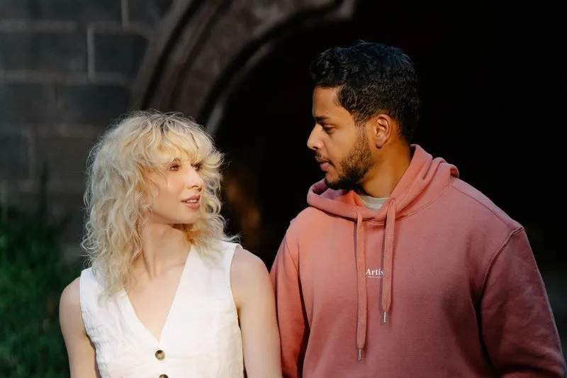A man and a woman look at each other as they talk quietly outdoors, in front of a stone wall