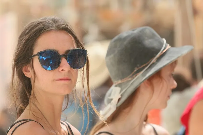Two young women in a sunlit festival crowd, one in sunglasses, the other in a wide-brimmed hat