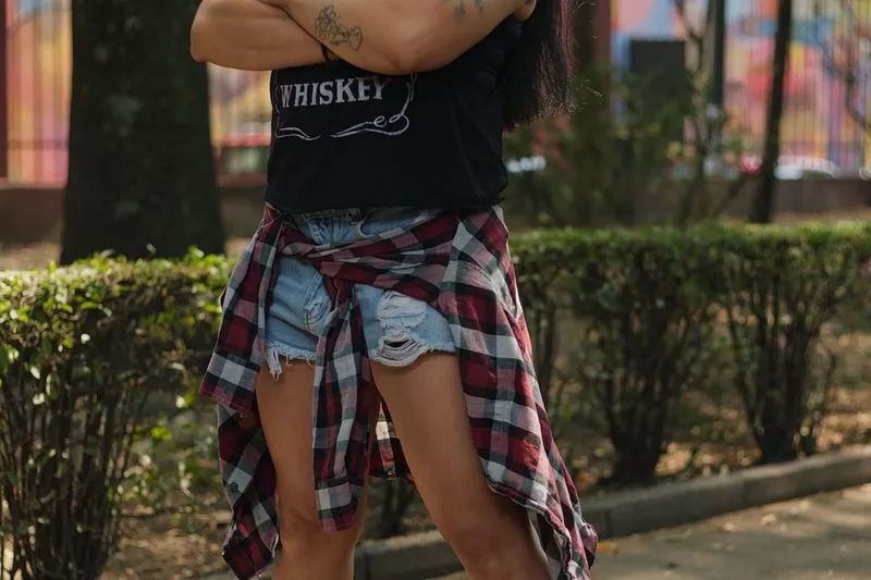 Young woman in a black tank top and ripped denim shorts, arms crossed, a red and black checked shirt tied around her waist