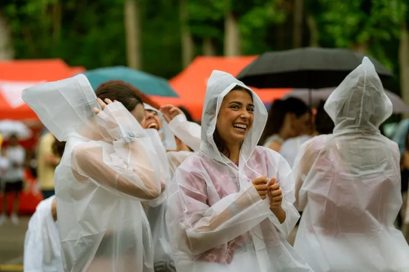 Group of festival-goers laughing under clear rain ponchos, in front of colourful tents, in the rain