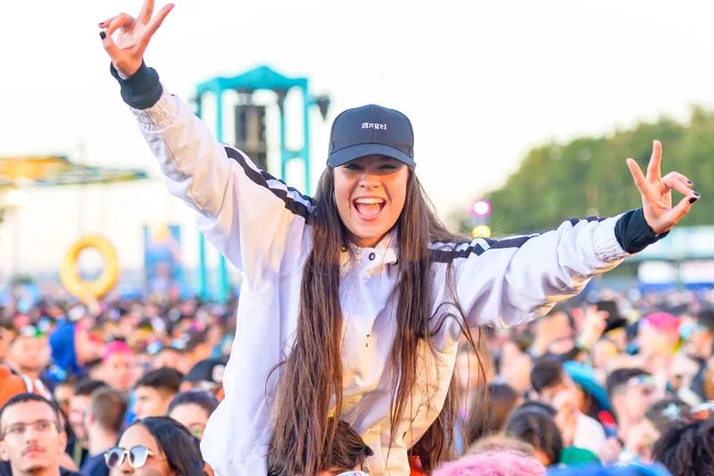 Young woman perched on a friend's shoulders, cap on her head and arms raised in peace signs, in the middle of a festival crowd at dusk