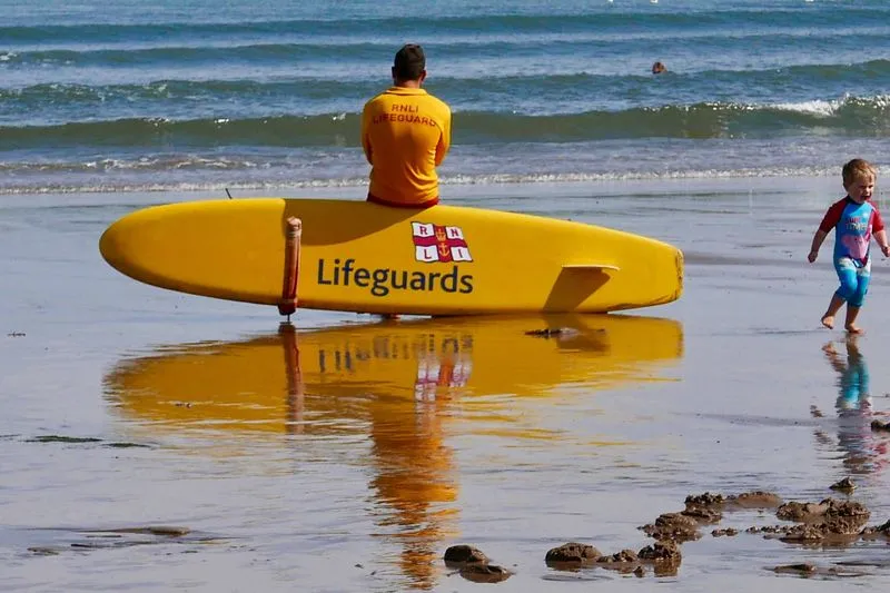 RNLI lifeguard sitting behind a yellow rescue board, watching the sea, as a toddler runs across the wet sand