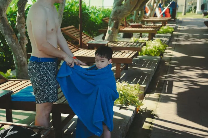 A father wraps his young son in a large blue towel beside poolside loungers