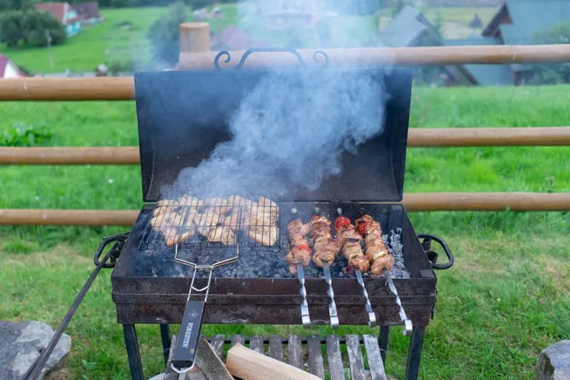 Rectangular charcoal barbecue with the lid raised, chicken pieces in a grill basket on one side and meat and tomato skewers on the other, smoke rising