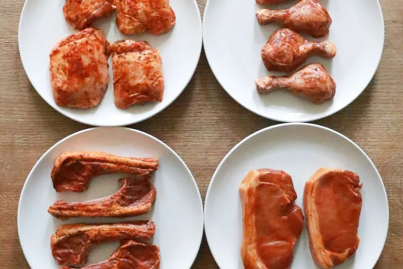 Four plates of raw marinated meat — chicken pieces, drumsticks, chops and pork steaks — coated in a spiced red marinade, on a wooden table
