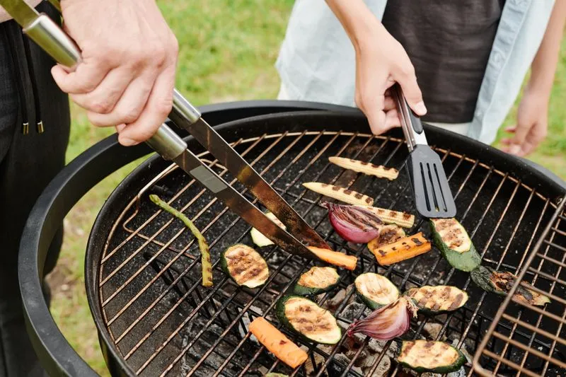 Small round barbecue, two people turning courgette, red onion and carrot slices on the grill with tongs