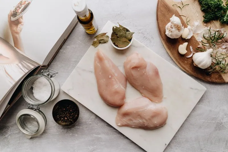 Raw chicken breasts on a marble board, next to a wooden board with garlic and fresh herbs