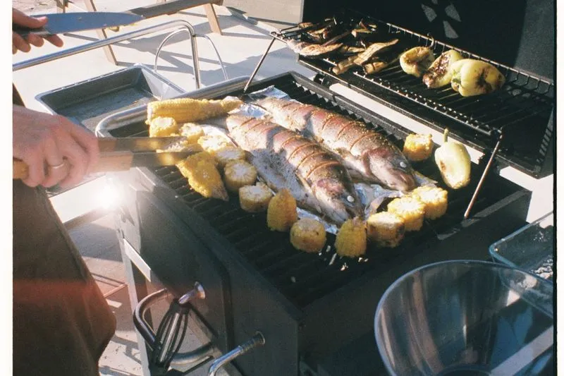 Several whole fish on a barbecue grill surrounded by corn on the cob, with peppers grilling on a nearby rack