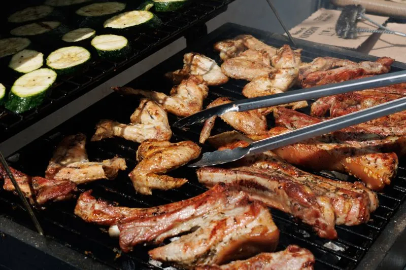 Courgette slices, chicken wings and pork ribs on a barbecue grill, with tongs turning the meat